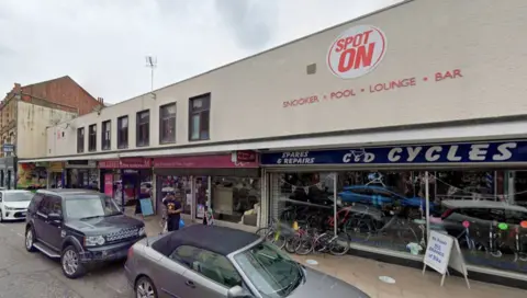 Several cars parked outside a two storey building with glass frontage featuring bicycles. Red lettering advertising snooker, pool and a bar is on the side