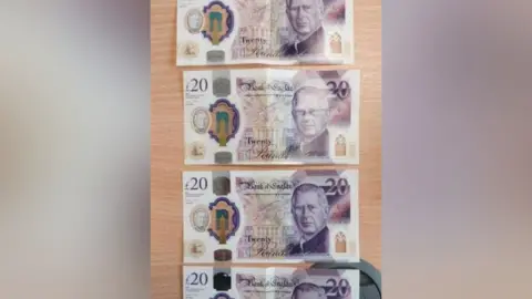 Four counterfeit £20 - bearing King Charles' image - laid out on a pine-coloured desk