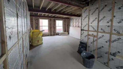 The interior of a building with a window on the far wall and wooden frames with plastic sheeting stretched between them on all walls. The wooden beams of the ceiling are exposed