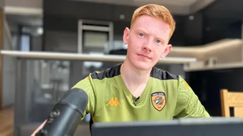 A 19-year-old man with short ginger hair wearing a green T-shirt with an orange Hull City badge on it. He is sitting at a desk behind a microphone and a computer.