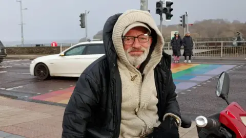 A man with a grey beard and glasses. He is wearing a black coat and cream jacket.