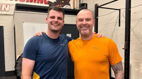 Wirral University Teaching Hospital Joe Clarkson, in a dark blue t-shirt, pictured with his arm around Grant Williams (right), in an orange t-shirt. There is gym equipment in the background.