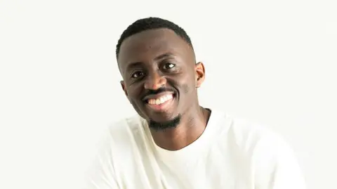 Joseph Charm is smiling at the camera while wearing a white crew neck top. He has dark hair and facial hair. He is in front of a white background.