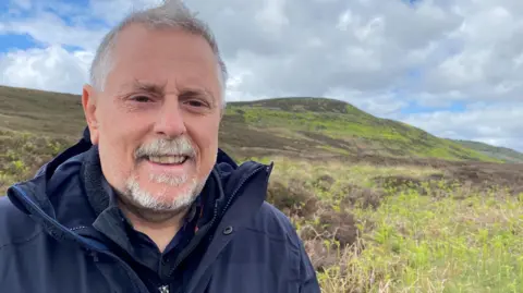 Mike Pratt is standing in front of the Simonside Hills. He is in his 50s and with grey hair and a grey moustache and beard. He is wearing a blue jacket and top