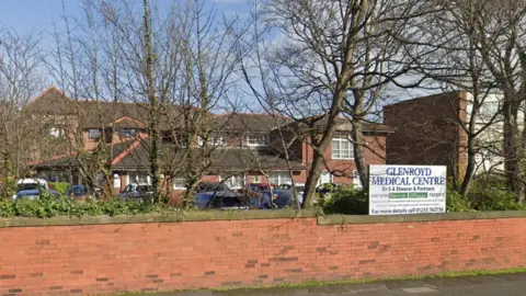 Google Street view image of Glenroyd Medical Centre. A complex of single storey and two-storey red brick buildings with cars parked in front. In front of them is a low red brick wall and many bare trees with a white sign advertising the practice