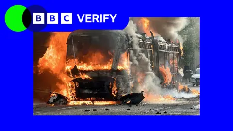 A bus on fire in Belfast, during a night of violence following a knife attack. There is blue and green BBC Verify branding in the top left corner.