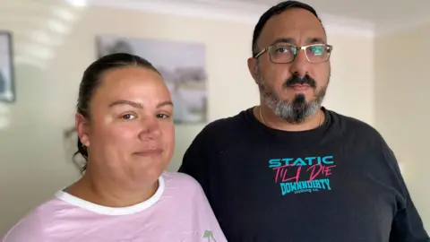 A woman and a man pictured in their home. The woman has dark hair tied into a ponytail and is wearing a pink t-shirt with a white neck. She is stood next to a man with short, dark hair and facial hair. He is wearing a black t-shirt with a blue and pink logo on the chest. Both of them are looking directly at the camera.
