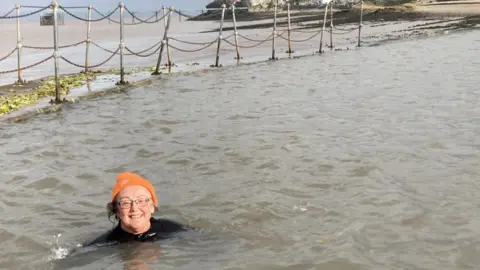 An older woman wearing a black wetsuit and an orange woolly hat, smiling to camera while enjoying a dip in the Clevedon Marine Lake.