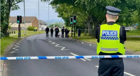 Brian Punter-Mathews/BBC Police line and officer on Middleton Ring Road in Beeston Park with another group of police officers in the distance investigating the site where a 19-year-old was hit by a car and killed on Friday.