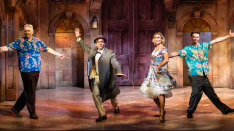 Jack Merriman Three male actors and one female are on stage lined up together in various dancing poses. Two have Hawaiian-style t-shirts and black trousers, one man has a trench coat, suit and a hat, and the woman has a frilly floral skirt with crinoline petticoats underneath. The stage setting behind is a large hall with floor-to-ceiling wooden doors in the middle