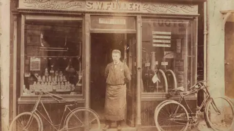 Family photo Sepia photograph of a man in a suit and lare apron standing in the doorway of S W Fuller shop. 