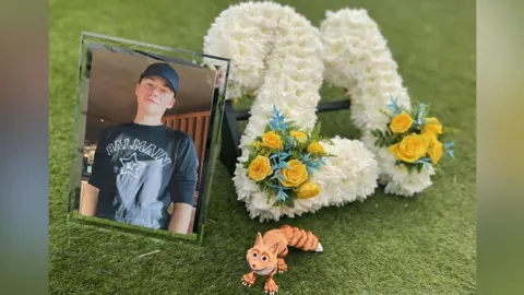A picture of Matthew Gray beside a floral display which says '21'. Matthew is wearing a black t-shirt and hat in the picture, while the floral display is white with two small bunches of yellow flowers.