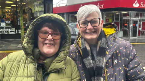 Anne and Jo standing in the high street. Anne is wearing a hooded grey coat, has short brown hair and is wearing glasses. Jo is wearing a purple coat with white, yellow and red polka dots, she has short white hair and is wearing glasses. 