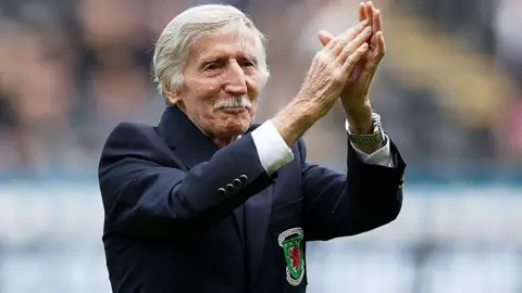 Cliff Jones, who has white short hair and a moustache, has his arms raised clapping. He has a blue blazer on with the Wales logo.