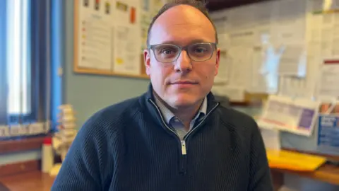 BBC/Charlotte Rose Dr Jamie Green wears grey glasses, and a blue zip up fleece over a blue checked shirt. He sits in his office with the walls covered in laminated notices.