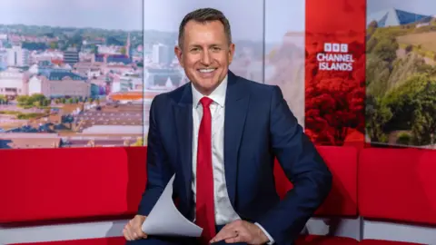 A man with short dark hair, smiling at the camera. He is wearing a navy suit, a white shirt and a red tie, and sitting on a red sofa in a TV studio. He has a set of white papers in his hand. Behind him is a backdrop showing the St Helier skyline, which says 'BBC Channel Islands' in white writing.