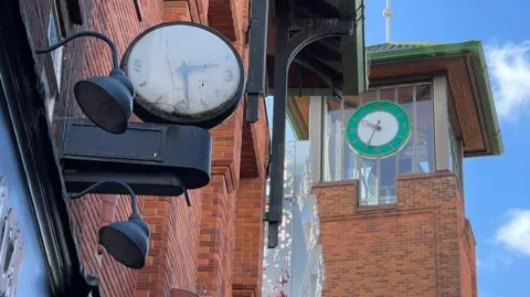A black clock with a white clock face attached to a shop building shows the time as 14:30. Behind it, a green and white clock is displaying the time as 10:35.