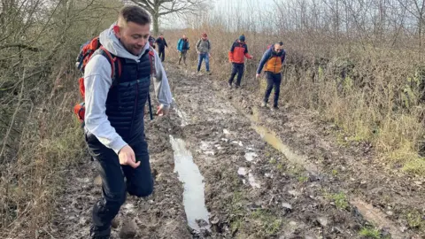 Mark Granger A man in the foreground photographed mid-leap through some muddy terrain with a string of other people behind negotiating their way through the mud