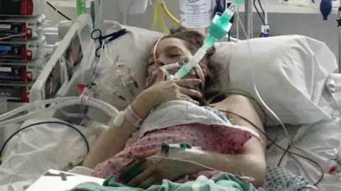 Helen holding baby Maeve. She has a large tube in her mouth and wires attached all over her body. There is a baby incubator to the left hand side