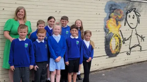 Bridge Farm Primary Children from Bridge Farm Primary school in Bristol smile at the camera. They are wearing blue jumpers and standing against a wall which has a piece of Banksy art on it - a child rolling a burning tyre