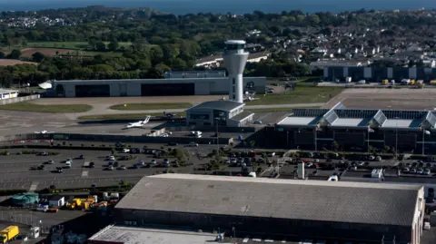 BBC Photo taken from an aircraft or tall building overlooking Jersey Airport. The airport's control tower is in the background. A car park with dozens of vehicles is in the middle of the image. Several hangars and other buildings are dotted around the site.