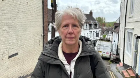 Charlie Stubbs / BBC News There is a woman with grey hair, a black unzipped overcoat, another white coat underneath that and a purple t-shirt. She is stood on the narrow street, with a white wall to her left and and steps to terraced houses to her right. There is also a white van behind her.