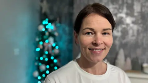 A woman with brown hair and a white t shirt smiles in front of a christmas tree 