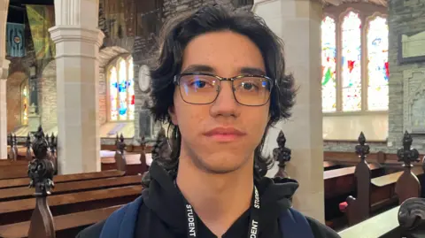 Luca has dark hair parted in the middle, curtains-style. He is wearing a black hoodie and around his neck is a lanyard with the word 'student' printed on it in white against black. 