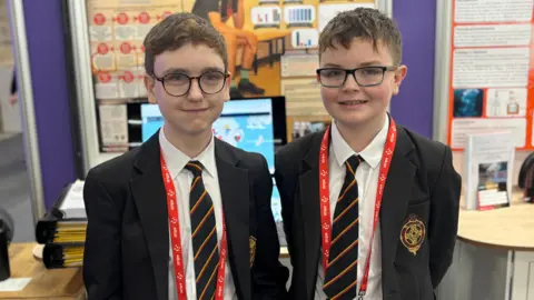 Two schoolboys. They both wear glasses. They are wearing a black uniform blazer with a white shirt and a tie with black, yellow and red stripes. 