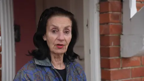 A woman wearing a purple jacket stands in the doorway of a brick building. She has shoulder-length black hair.