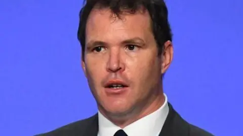 Lee Waters in a dark jacket and tie against a blue background 