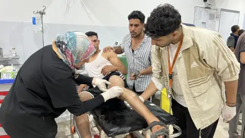 Getty Images An injured man is lying on a hospital trolley while medics treat his wounds, another man is looking on concerned