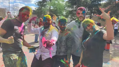 A group of five young people covered in colourful powder pose and smile. 