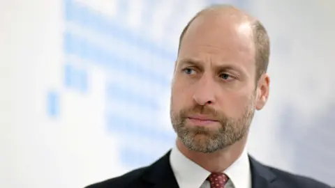Prince William, wearing a black suit, white shirt and red tie