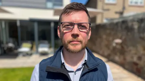 Josh King, in a blue body warmer, with a brown beard and hair, wearing black glasses. He is standing in a residential garden.