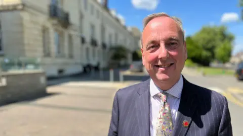 Ian Thorn smiles at the camera wearing a pinstripe suit and floral tie. Behind him, the council HQ is out of focus, a sunny day