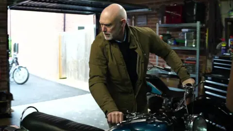 A man in a garage looks down to his right as he has both hands on the handlebars of a motorcycle. 