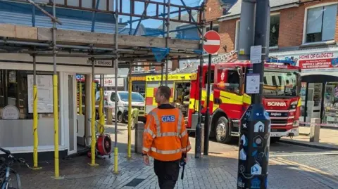 A man is standing on the corner of a street which has a fire engine in the middle of a road. To the left is a building with metal scaffolding around it. 