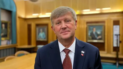 Peter Harris looking at the camera wearing a suit and tie in a grand meeting room which is out of focus in the background.