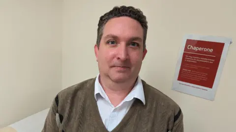 Dr Philip Hine in a doctor's room, wearing a white shirt and brown jumper. There is a red sign on the wall behind him offering a chaperone service to patients.