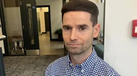 A man with brown hair wearing a checked blue and white shirt. He is sitting in an office.