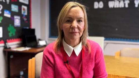 Orlagh Barrett is sitting on a chair and is staring at the camera. She has shoulder length blonde hair and is wearing a pink jumper with a white collar. She has a red lanyard around her neck. In the background is a desk and a classroom noticeboard.