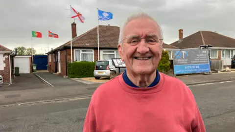 A man in his 80s is wearing a pink jumper, standing outside of his house which has four flags flying above it. 