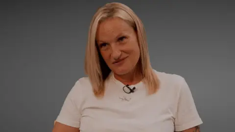 Cheri Curran, sitting down, taking part in a film, looking off to the camera, she is wearing a white T-shirt, with a microphone at her neck, and a necklace round her neck. She has long straight blonde hair. 