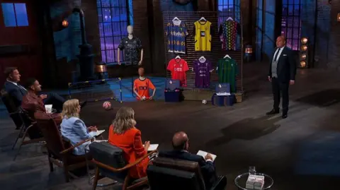 Image of Alex Street inside the Den. A wider shot showing a selection of different football shirts hung. Dragons Peter Jones, Steven Bartlett, Deborah Meaden, Sara Davies and Touker Suleyman look on. 