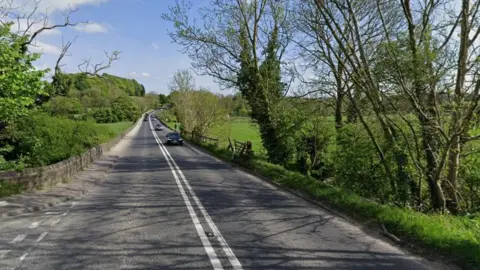 A street view image of a road winding its way through the countryside. Three cars can be seen travelling along the right lane, with trees and shrubs running along either side of the road.