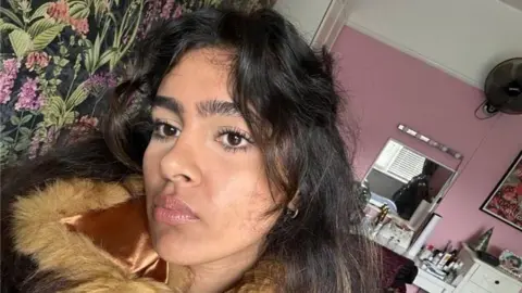 Mashaal Chughtai A young woman with long dark wavy hair and a fringe takes a selfie in a bedroom with pink walls and floral wallpaper. She wears a brown fur-collared jacket and looks directly into the camera with a serious expression. Behind her is a white dressing table with a mirror and various items on top.