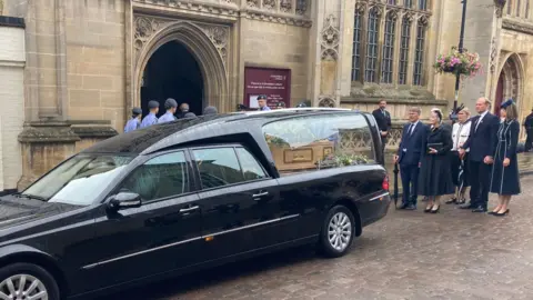Luke Deal/BBC A black hearse is parked outside St Edmundsbury Cathedral. A coffin is visible inside with flowers on it. Behind the hearse are family members of Lord Tebbit.