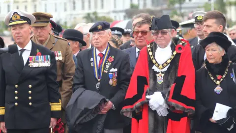 Tony Mottram A man dressed as an admiral stand solemnly. He wears a white hat, navy jacket, white shirt and tie. On his chest are a series of medals hung by colourful ribbons and at his side hangs the shinning gold handle of a sword. 