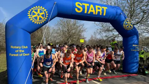 A large group of runners start running at the start line of a half marathon. The start line is a blue inflatable arch with a yellow logo for the Rotary Club of the Forest of Dean, and there is a red line on the floor. The group of runners is mostly composed of men in tank tops, shorts and trainers.